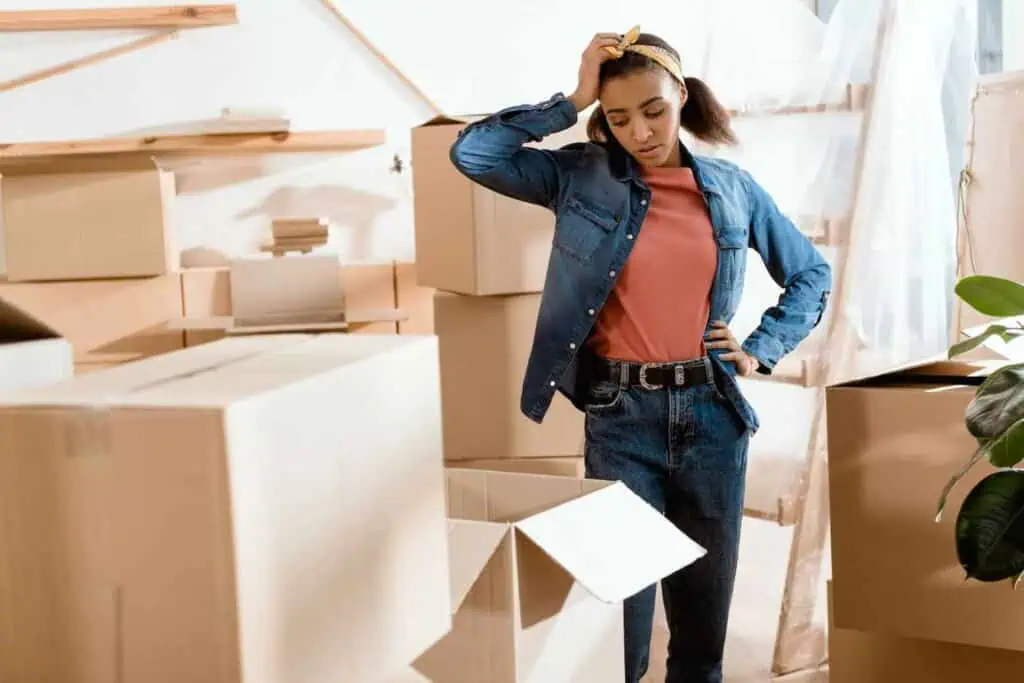 A woman with an upset and stress expression stands in the middle of stacks of cardboard. The one directly in front of her is open 