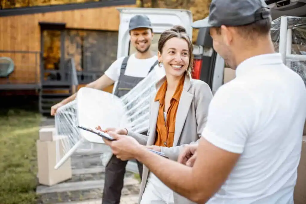 a woman in a grey pantsuit chats happily with her movers who are taking things out of a moving truck