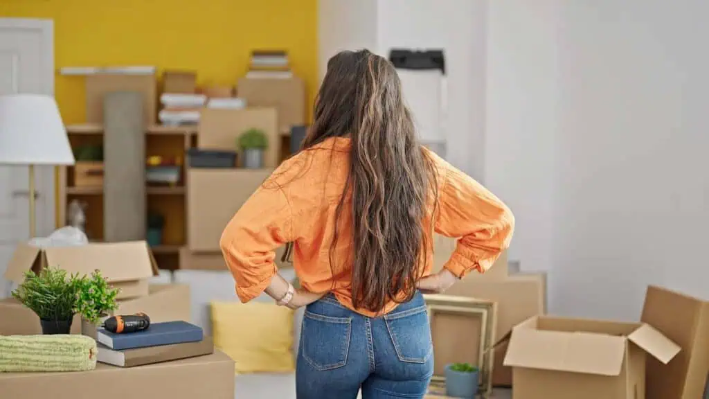 A woman with her hands on her hips stands with her back to the camera looking over a room filled with boxes and furniture 