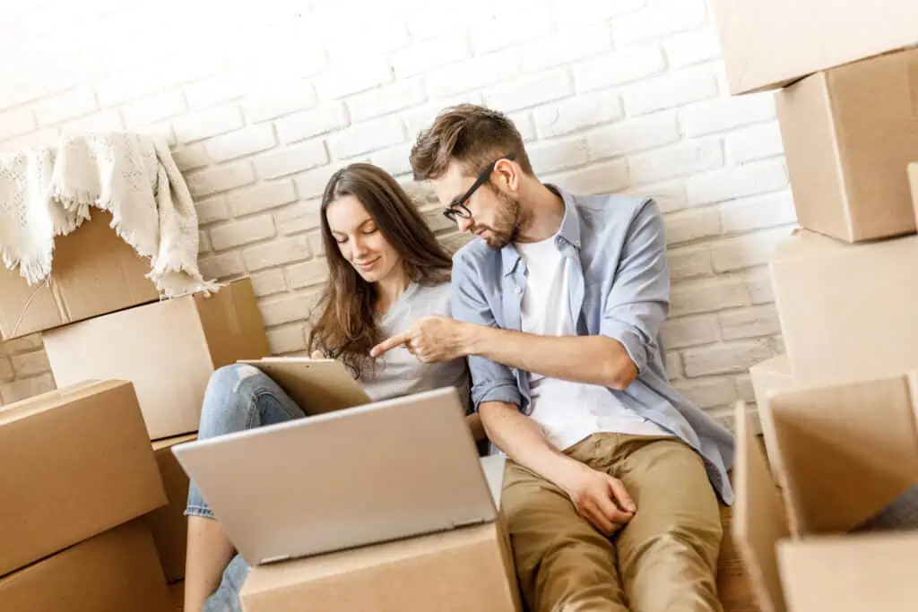 a couple surrounded by moving boxes discusses something in front of a laptop