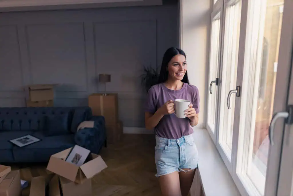 a woman stands by a sunny window holding a cup of coffee. A half-open moving box is behind her.