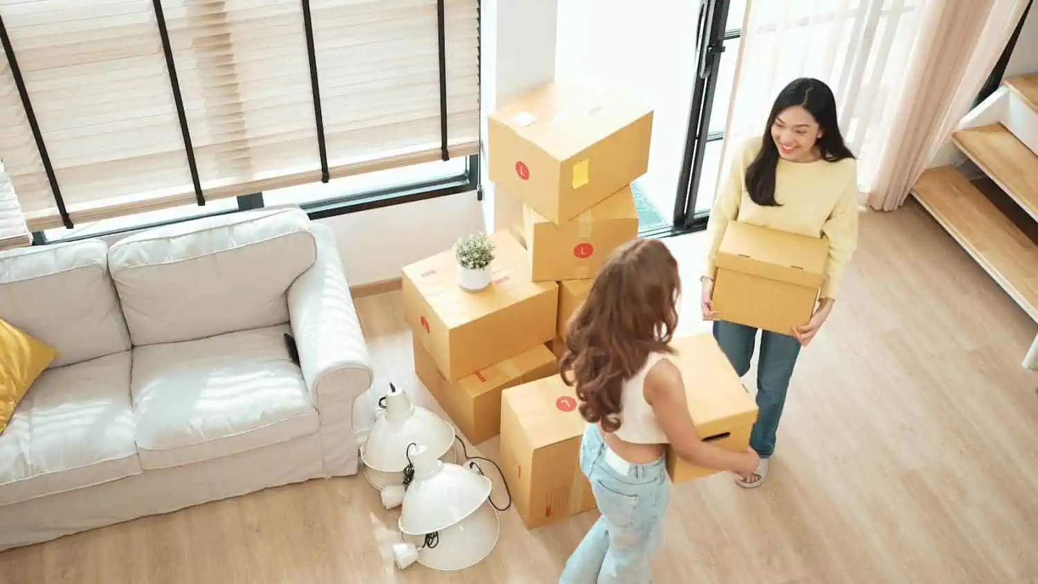 Two women carrying boxes into a home. There is a pile of more boxes next to them.