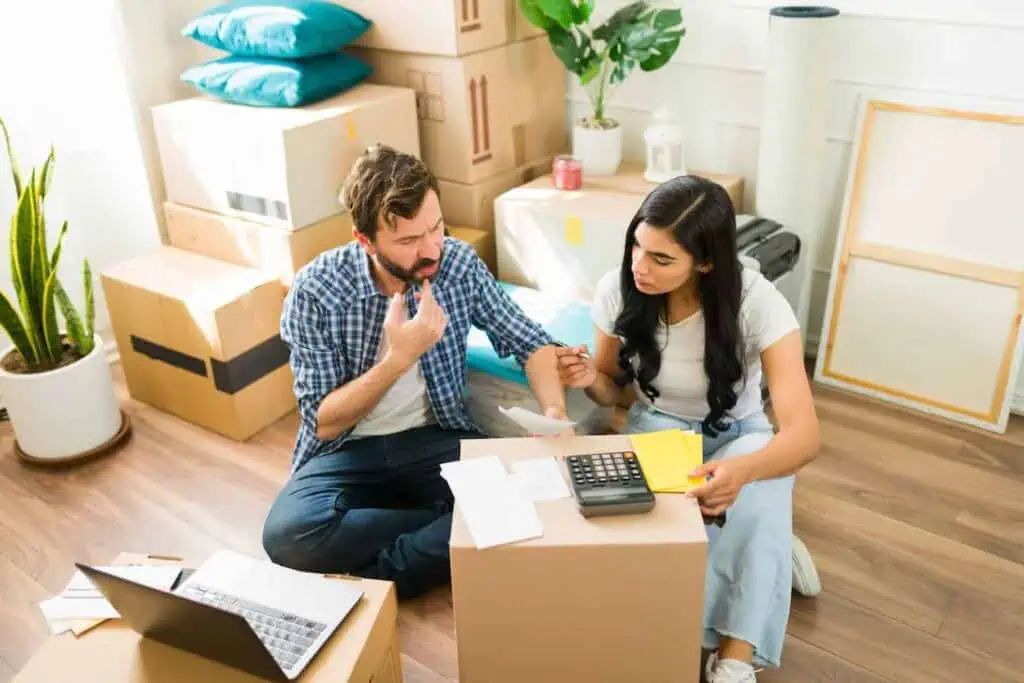 A couple in their home that's partially packed up looks contemplatively at a calculator and documents