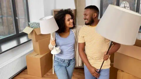 A couple carrying lamps in a room filled with boxes smile at each other