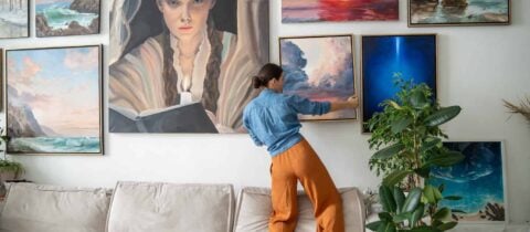 a woman takes down an oil painting from a tall wall that has many more covering its surface