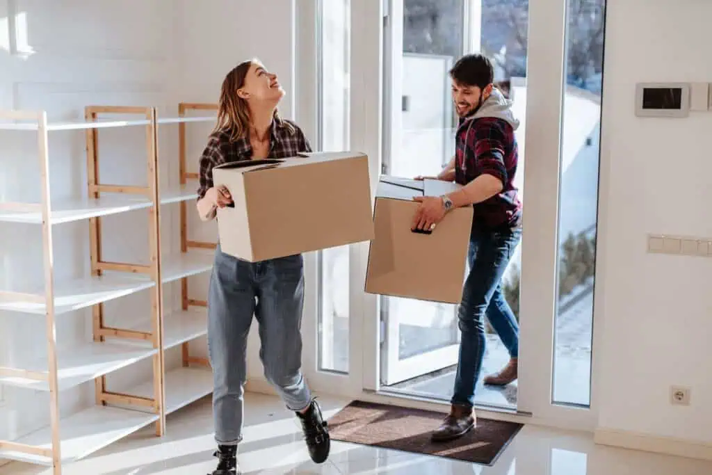 A man and a woman walk into their new home happily while carrying boxes