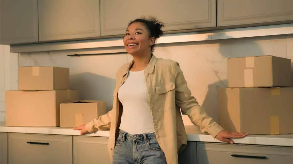 A woman leans back, relieved and happy, against her kitchen counter. There are several boxes on the countertop behind her.
