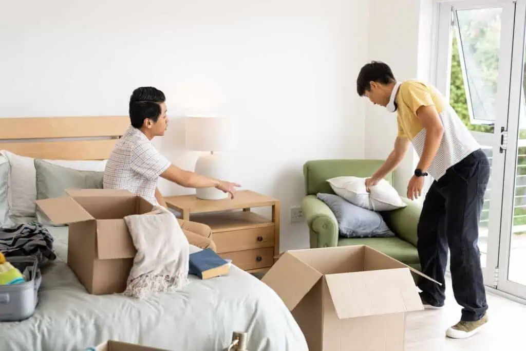One man sitting on a bed instructs another to pick up a pillow from a nearby chair. There are several boxes already packed around them.