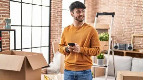 a man stands in a room full of moving boxes while holding his smart phone