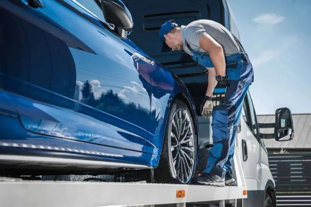 A worker in overalls loads a blue car onto a truck trailer for transport