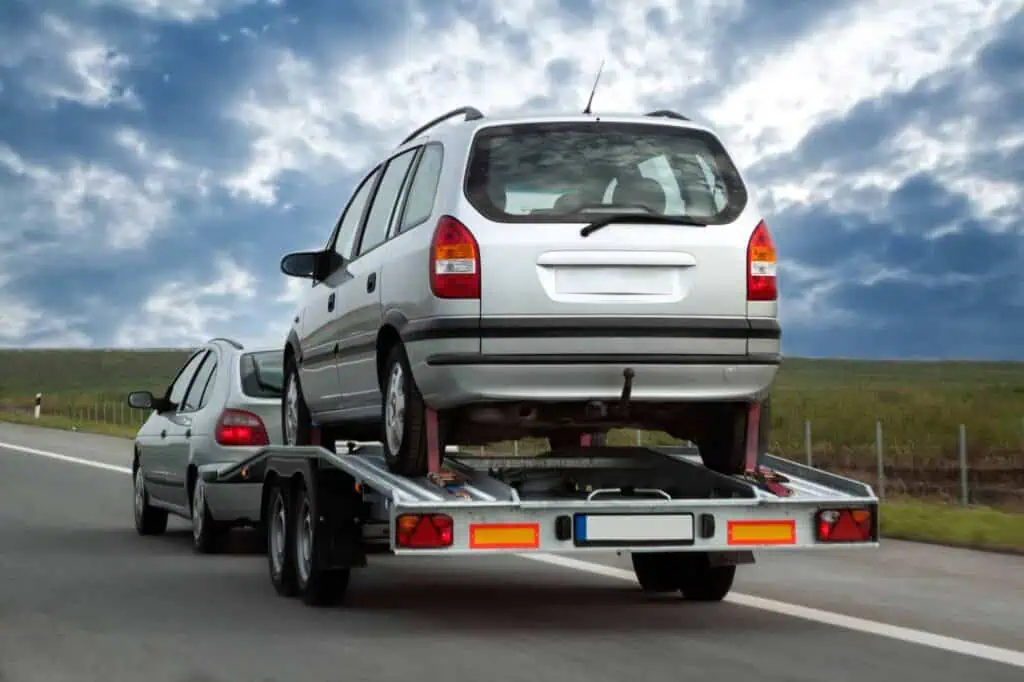 A car towing a trailer with another car attached to it. Both are traveling down a country highway.