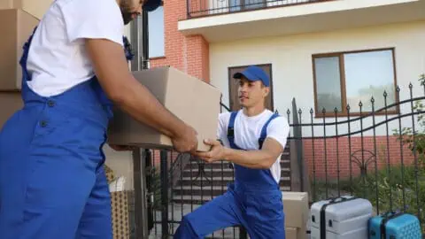 two male movers in blue overalls help unload moving truck full of boxes and furniture