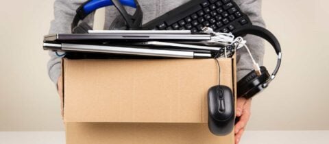 a person holds a box full of electronic waste, which include laptops, a keyboard, headphones, and a computer mouse