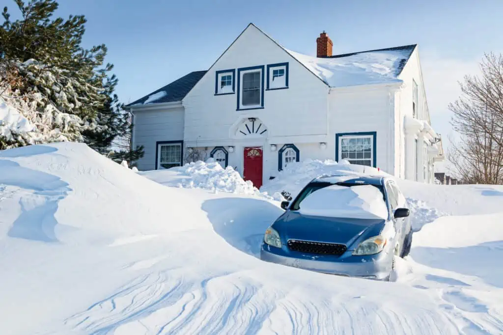 a car trapped in a snowed-in driveway in front of a white and red home. There is more snow piled up in drifts on either side of the lawn.