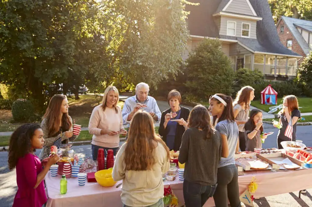 neighbors talk around a table during a block party. they're of all different ages, showing a mix of different families.