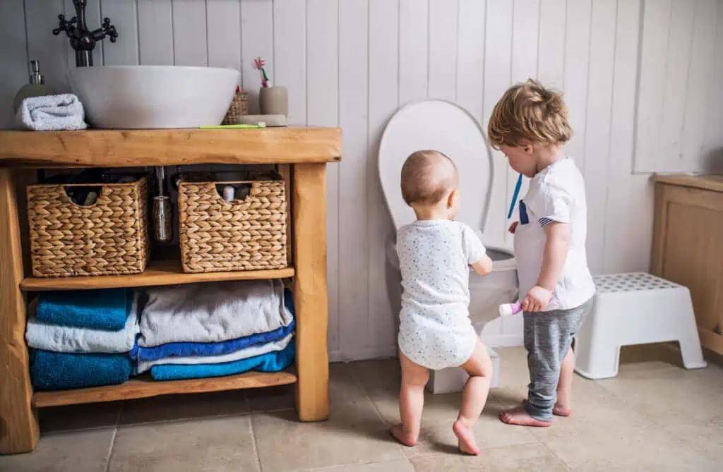 two toddlers standing by a toilet in a bathroom. There is a counter with a sink on top and blue towels folded below to the left. 
