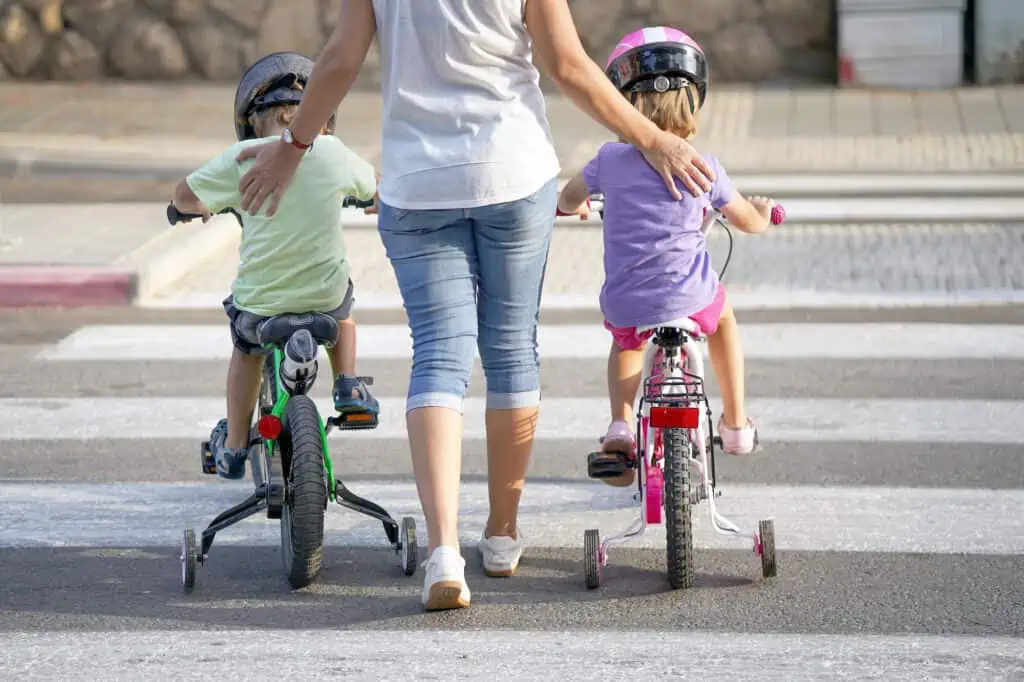 a woman guides two children riding bikes with training wheels across a crosswalk