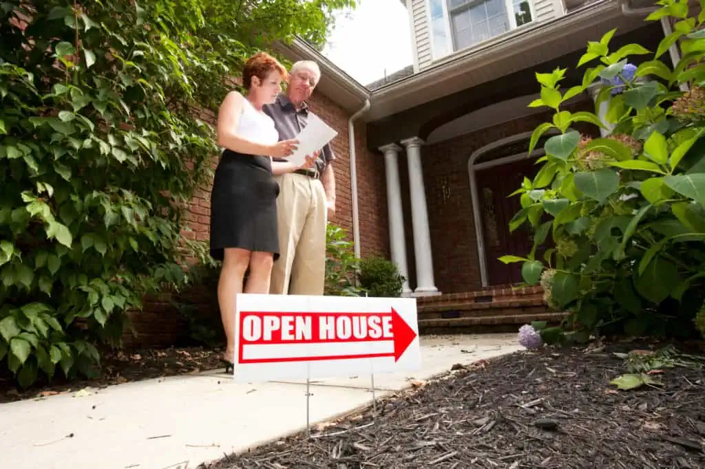 a senior couple stands on a walkway in front of a brick home. There are bushes to either side, and in the foreground is a sign saying "open house"