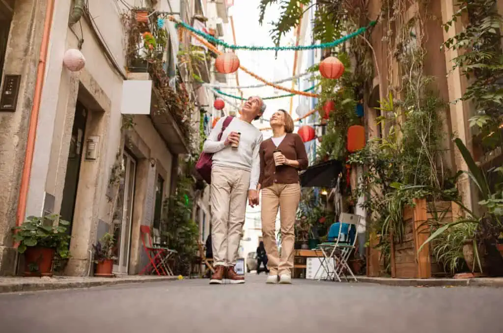 a retired couple walks along a street lined with plants, colorful lanterns, and banners hung between the houses on each side of the road