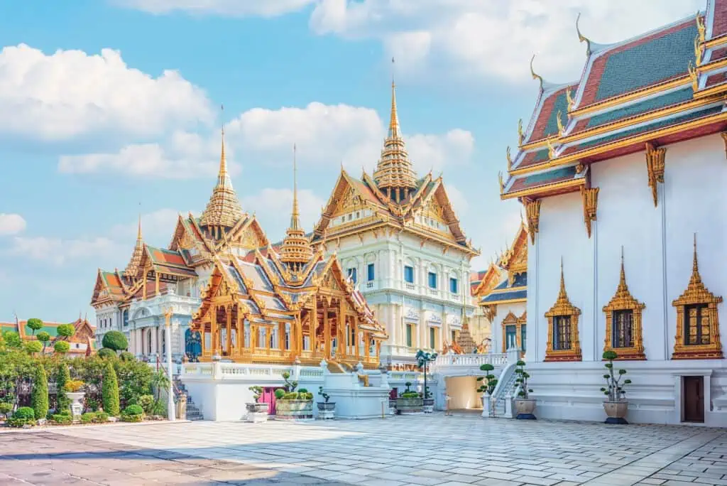 The Grand Palace in Bangkok, which is made up of white-faced buildings with golden roofs, and many multicolored accents. The photo is taken from an alley leading between these buildings