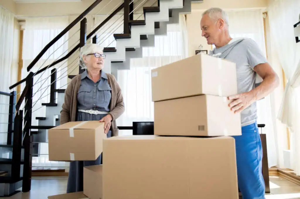 a senior couple stands in front of a stairwell, surrounded by boxes they've packed up for their retirement abroad