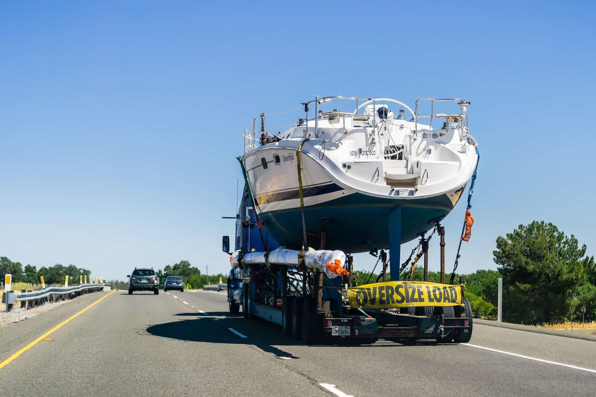 a truck transports a large boat along an interstate. There is a yellow banner hung along the trailer that reads Oversized Load