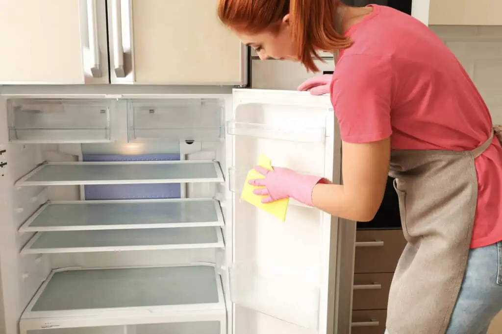 a woman wearing pink rubber gloves uses a sponge to clean out the interior of a fridge
