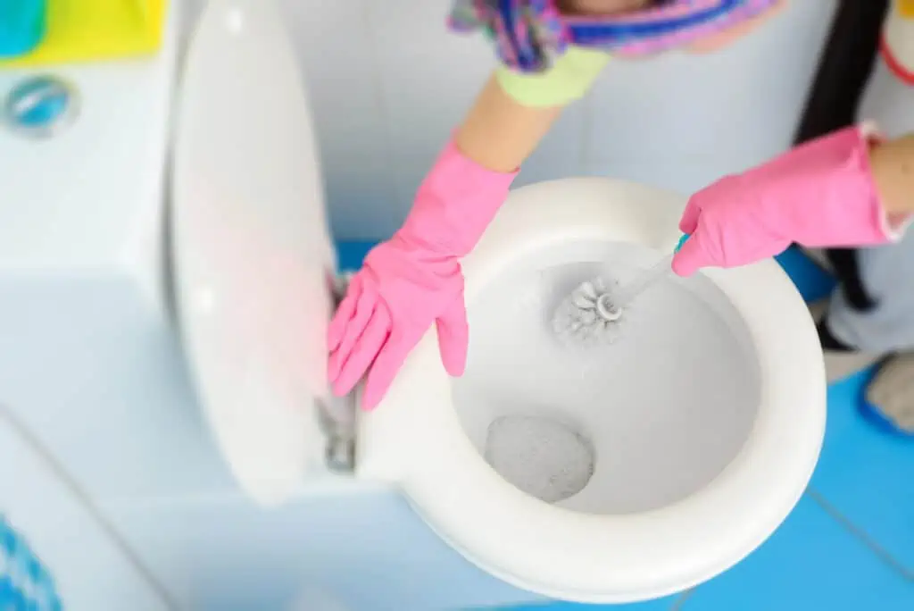 a woman scrubs under the lip of a toilet bowl with a bristled brush