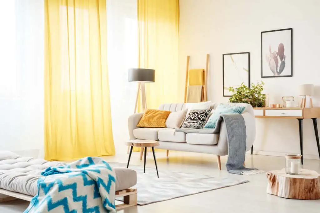 a view of a living room. Natural light is flowing in from large windows with semi-transparent yellow and white curtains. There is a white couch with multicolored throw pillows, as well as a settee and turquoise blanket in the foreground