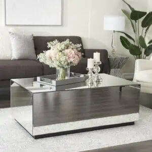 a solid square mirrored coffee table in the middle of a living room. The reflection of the floor and the flowers and candles on top are visible in the table's mirrored surface