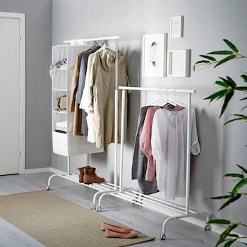 a two white clothing racks set up by a wall. One is taller and contains a set of mesh shelving for shoes and handbags. The other is shorter and has blouses hanging from it. 