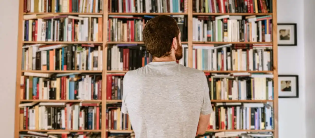 a man stands contemplatively in front of a full bookshelf that takes up one wall of a room