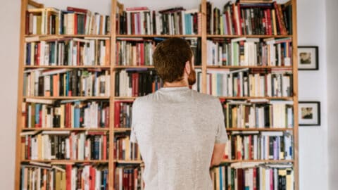 a man stands contemplatively in front of a full bookshelf that takes up one wall of a room