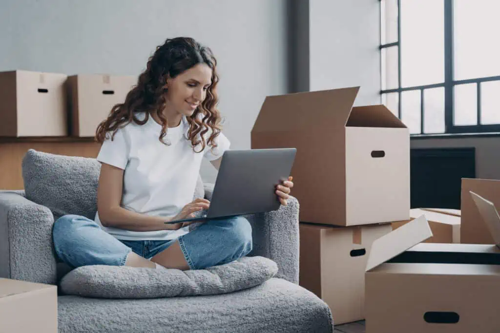 a woman sits on a chair with a laptop. She's surrounded by packed boxes as she plans a move