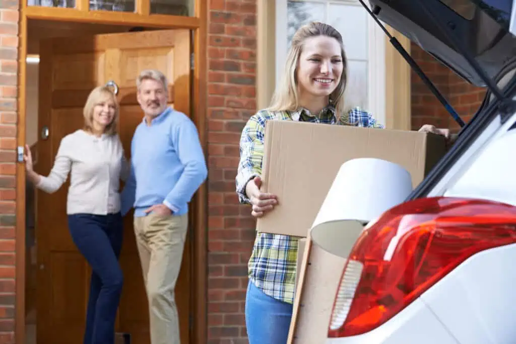 a young adult woman packs the back of her car with cardboard boxes while her parents watch from the entrance to their home
