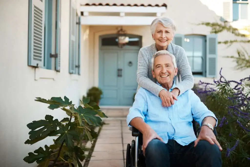 an elderly couple poses in front of a house with blue doors and eaves. The man is sitting in wheelchair. 