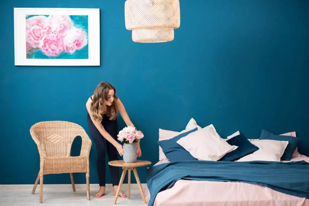 a woman places. vase of flowers on a table in a staged room. The wall is a shade of deep blue and so are the sheets on a bed nearby. 