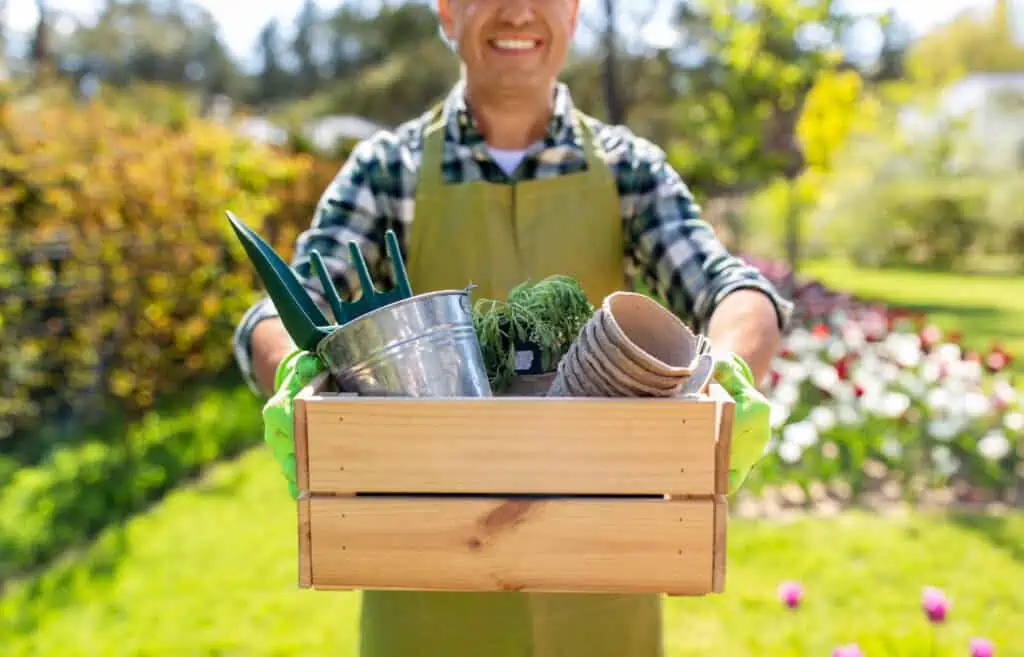 a man carries some of the tools needed to move a greenhouse, like a small spade and bucket, in a wooden box.