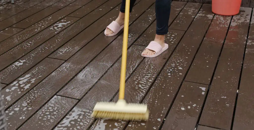 a person scrubs a wooden floor with a long-handled brush, water, and cleaner