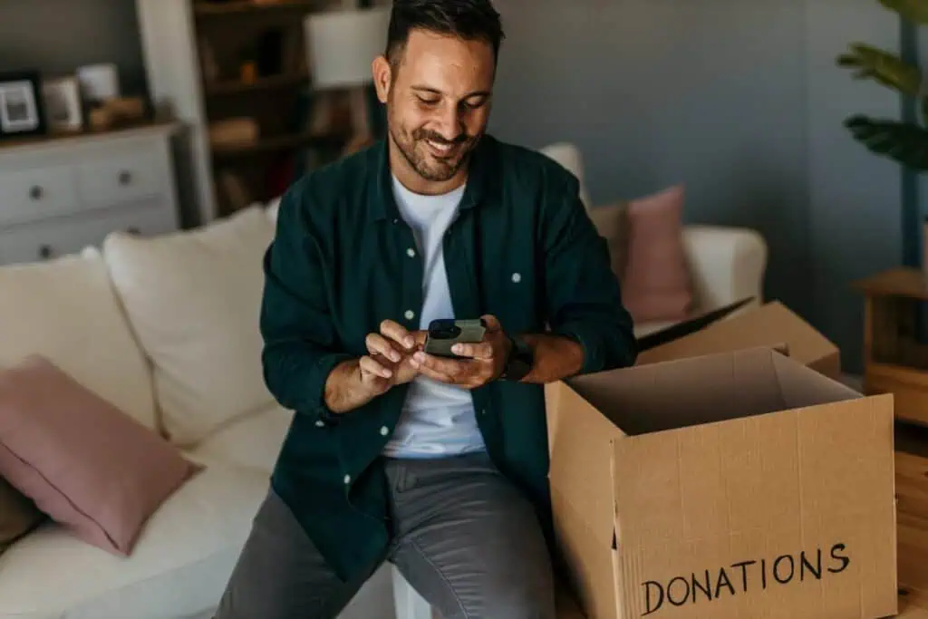 A man cheerfully looks at his phone. He's on a couch next to a box of items set aside for donation