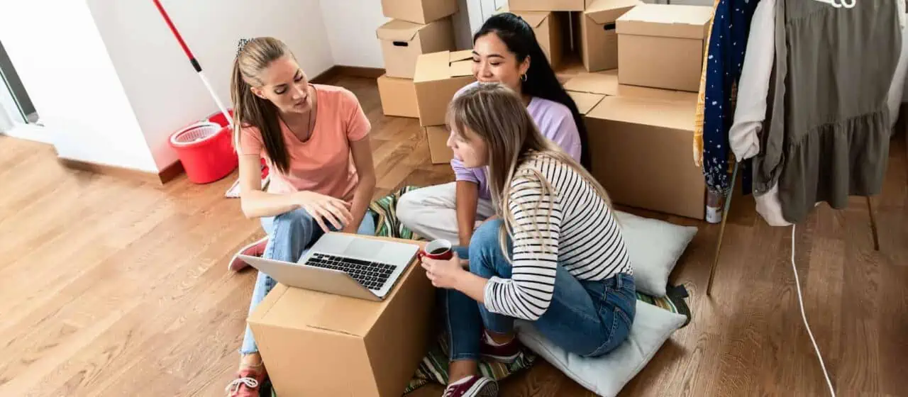 three young women sit around a computer in a room full of cardboard boxes