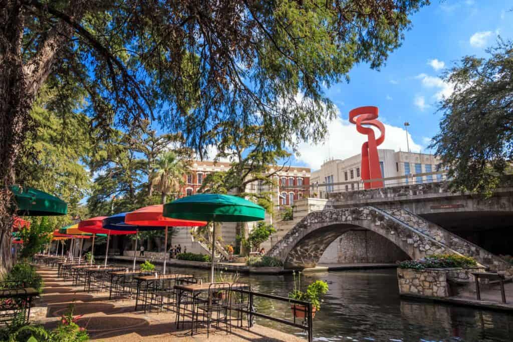 A view of San Antonio's river walk. There is a set of colorful umbrellas over tables and a bridge in the photo. 