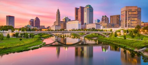 A view of downtown Columbus, OH at sunset. The buildings and skyscrapers border a river and bridge.