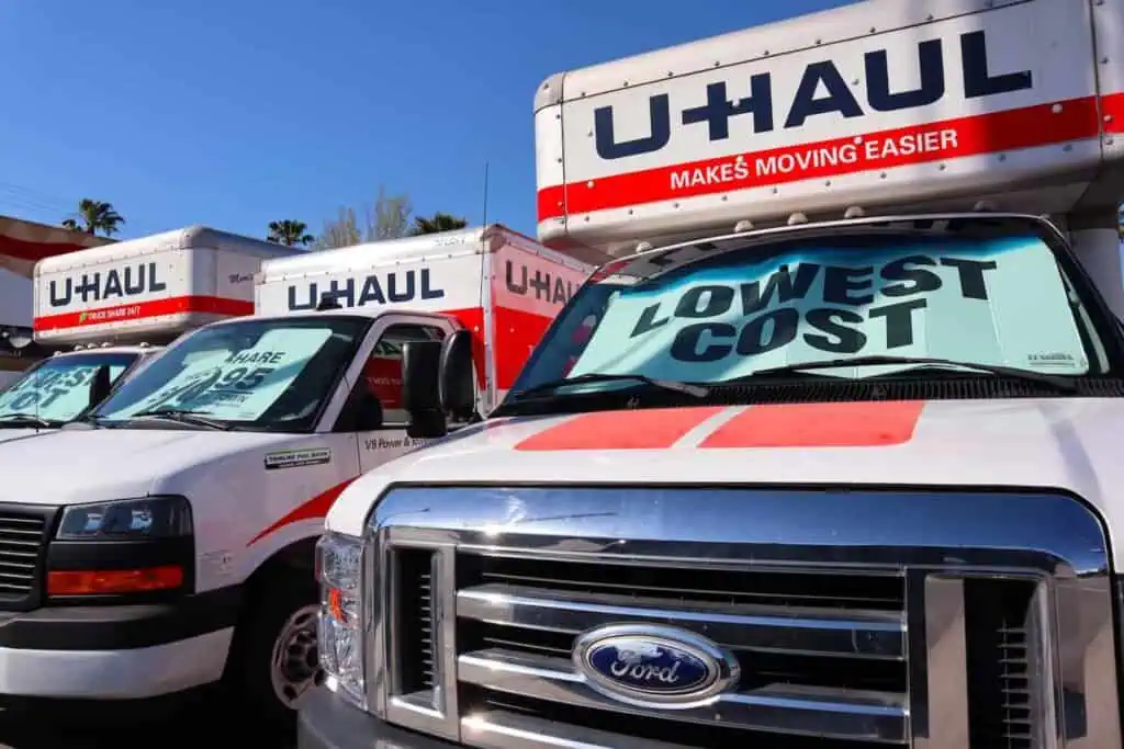 several uhaul trucks are parked in a row. There is a shade screen in their windows that reads "lowest cost"