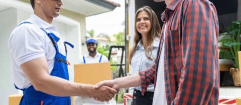 A couple shakes the hand of one mover while others move boxes out of a truck in the background