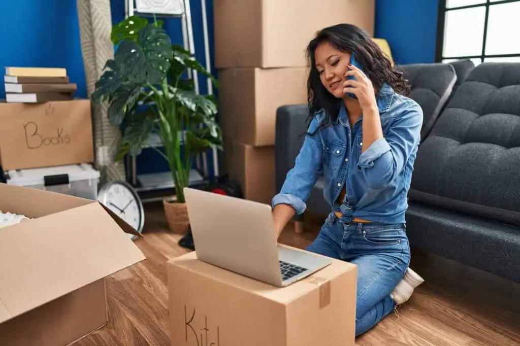 A woman sits on the floor in a room filled with boxes. She's on the phone while typing on a laptop. 