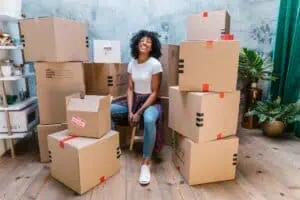 A smiling woman is sitting on a stack of boxes in an empty room, surrounded by piled boxes