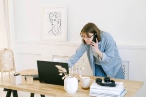 A woman stands before a desk, speaking on the phone while working on her laptop