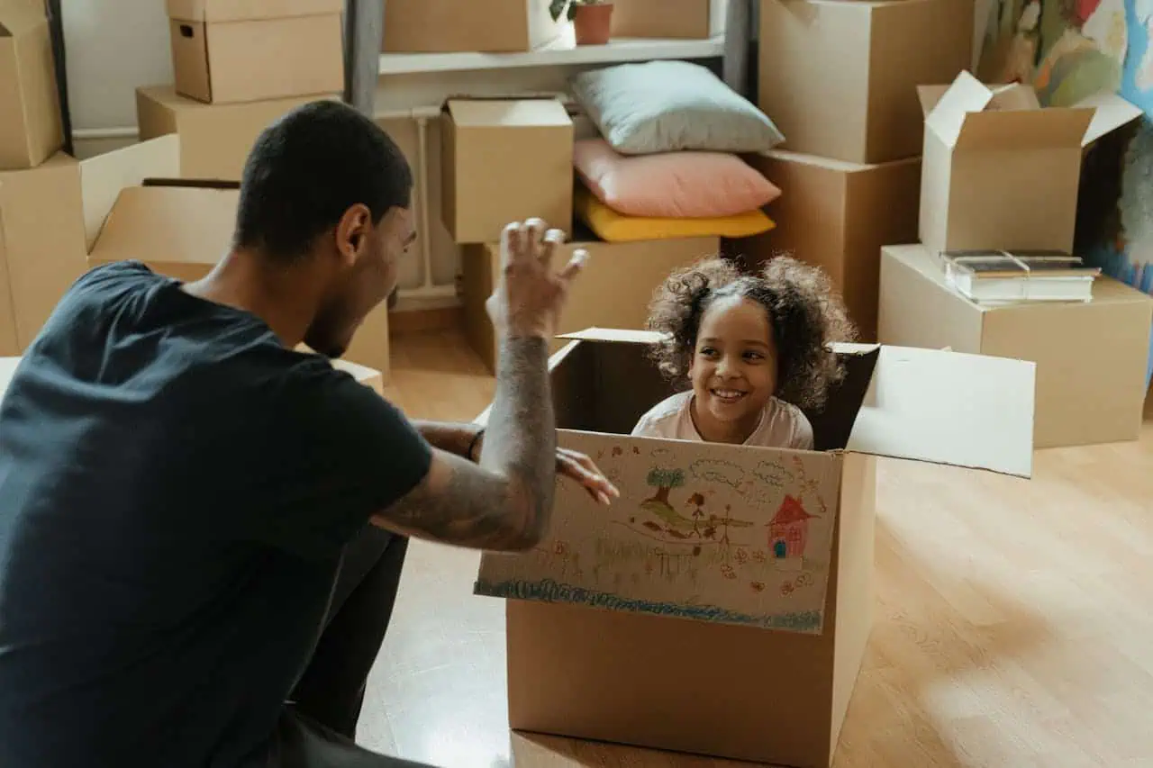 A man plays with a smiling child sitting in a decorated cardboard box surrounded by other boxes, indicating a moving process.
