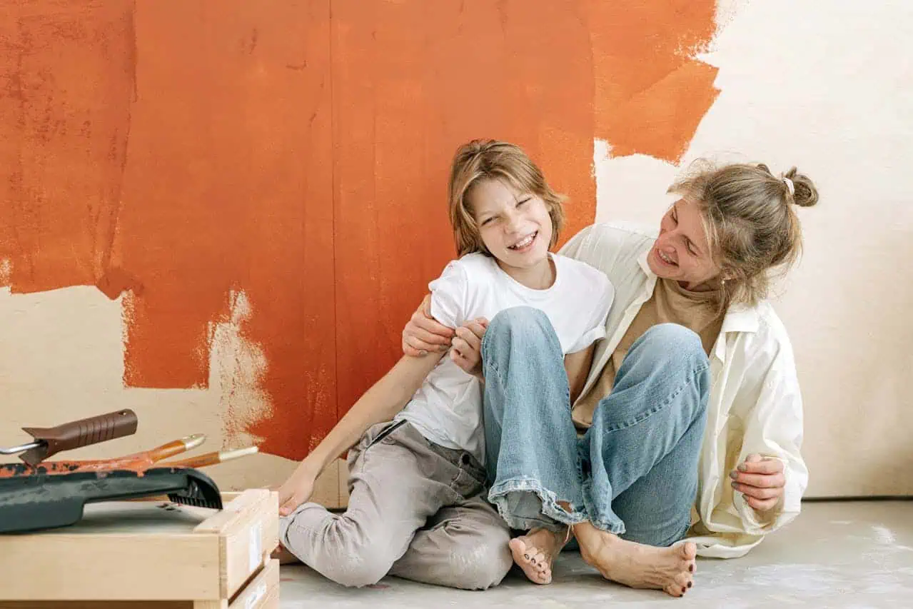 A woman and a child sit on the floor, smiling, in front of a vibrant orange-painted wall, indicating they've been painting it